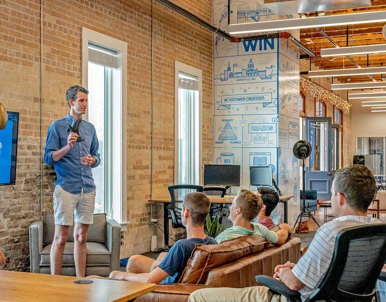 A number of people in a room that appears to be a creative office. One of the people is standing and talking. The others are sitting comfortably. 