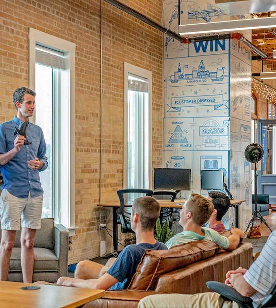 A number of people in a room that appears to be a creative office. One of the people is standing and talking. The others are sitting comfortably. 